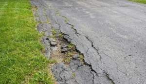 Deteriorated asphalt edges with large cracks and grass growing through the broken pavement.