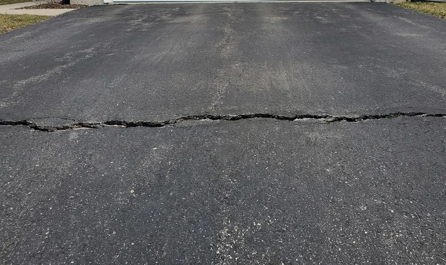 A wide transverse crack running horizontally across the width of an aging asphalt driveway pavement.