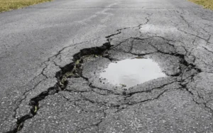 A large, deep pothole filled with standing water surrounded by severely cracked and sinking asphalt pavement.