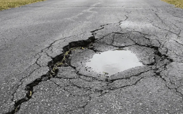 A large, deep pothole filled with standing water surrounded by severely cracked and sinking asphalt pavement.