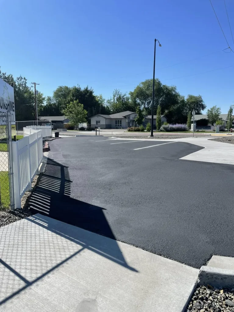 A bright daytime view of a newly sealcoated black asphalt parking lot featuring crisp white parking stall lines. Located next to a white fence and a concrete sidewalk, this complete asphalt maintenance and striping project was finished by Select Coatings in the Treasure Valley.