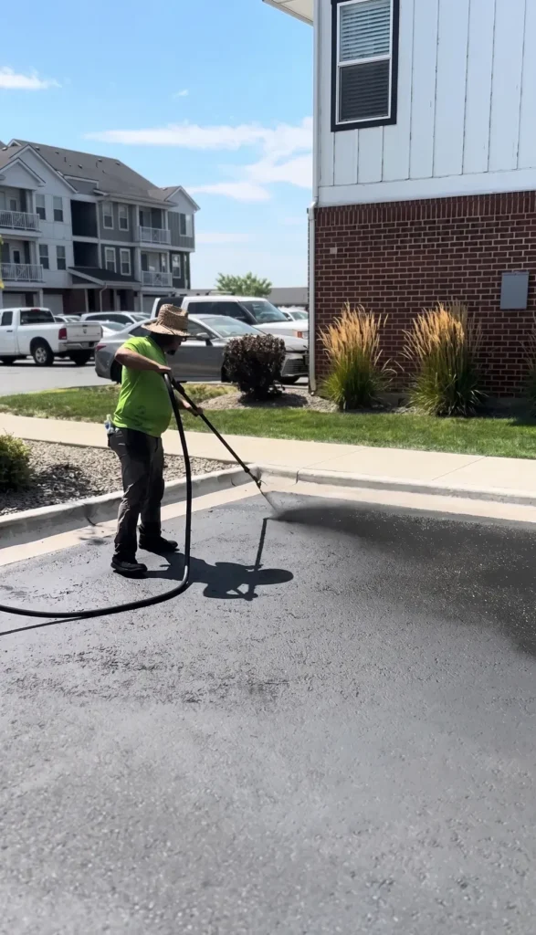 A professional technician from the Select Coatings team applies a protective asphalt sealcoat to a residential parking lot in Treasure Valley, Idaho. The worker, wearing a high-visibility green shirt and a straw hat, uses a precision spray wand to evenly coat the pavement near a red brick building. The high-quality sealant provides a dark, refreshed finish and long-lasting protection against the elements.